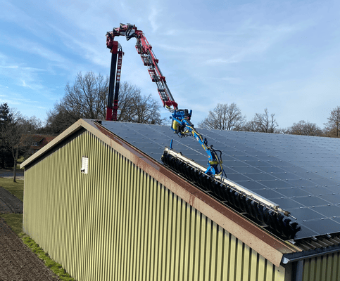 LKW mit Kran reinigt Solarzellen auf grünem Dach; klarer Himmel im Hintergrund.