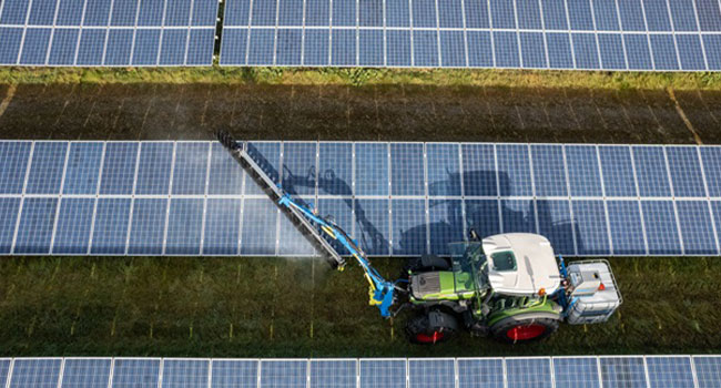 Traktor sprüht zwischen Solarmodulen auf grünen Feldern.